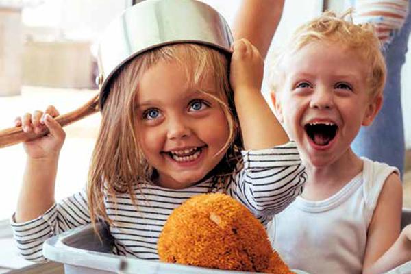 zwei lachende Kinder mit Kochtopf auf Kopf sitzend im Wäschekorb